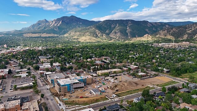 Aerial view of Boulder with the Flatirons in the background, showing urban development, construction sites, and lush green trees under a blue sky in Boulder, Colorado, United States.
