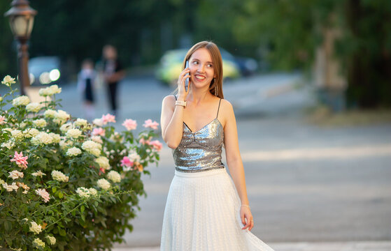Young smiling woman talking on smartphone while walking near blooming roses in a city park on a summer evening. Happy lifestyle, communication, technology and outdoor leisure.