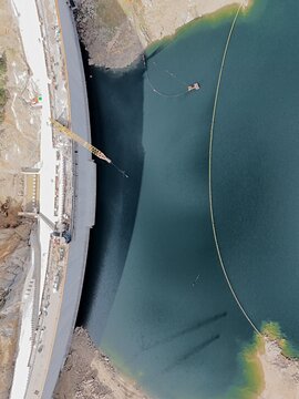 Aerial view of Gross Reservoir dam construction with a large crane, concrete structures, and a floating boom on the water in Boulder County, Colorado, United States.