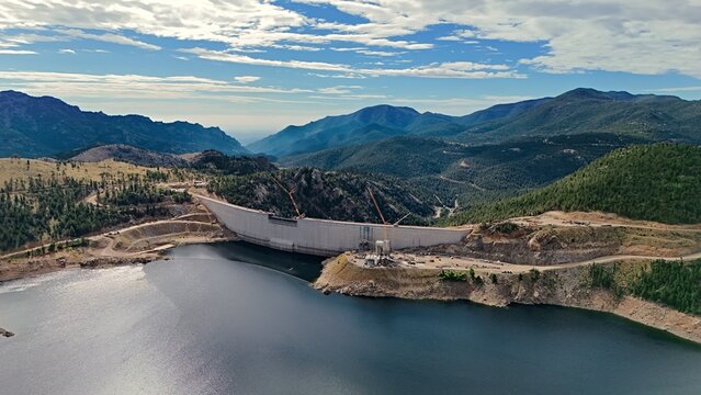 Aerial view of Gross Reservoir dam with construction cranes and concrete structure surrounded by forested mountains and reservoir water in Boulder County, Colorado, United States.