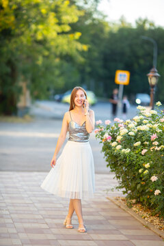 Young smiling woman talking on smartphone while walking near blooming roses in a city park on a summer evening. Happy lifestyle, communication, technology and outdoor leisure.