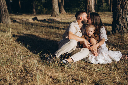 Family of three embracing outdoors in sunny summer weather, parents kissing while sitting with child in forest
