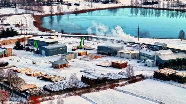 Aerial view of a snowy lumber yard with stacks of timber and industrial buildings near a frozen lake in Latvia.