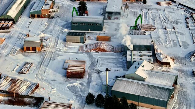 Aerial view of a snow-covered lumber mill with stacks of logs and industrial buildings in Latvia.