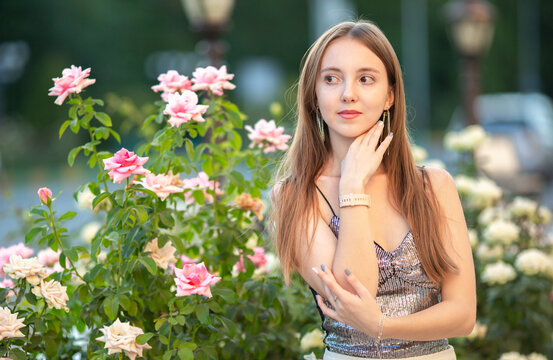 Elegant young woman posing among blooming pink roses in a summer garden. Romantic beauty portrait with long hair, stylish outfit and natural floral background, lifestyle concept.