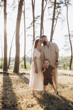 Parents hold their child's hands, smiling while walking through a pine forest on a warm summer day