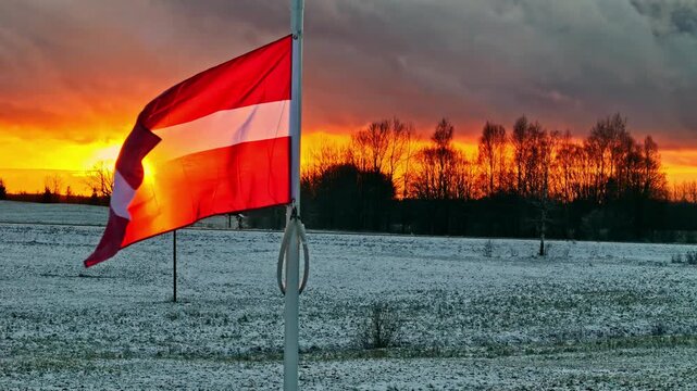 Aerial view of the flag of Latvia waving on a pole over a snow-covered field during a vibrant sunset in Latvia.