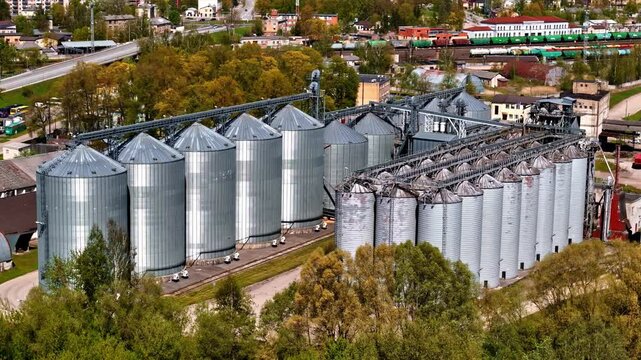 Aerial view of a large industrial grain silo facility with metallic storage tanks surrounded by trees and railway tracks in Riga, Latvia.
