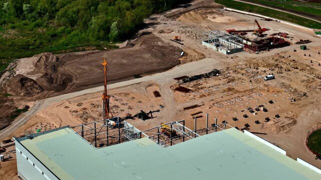 Aerial view of a large construction site in Riga, Latvia, featuring steel building frames, a tall drilling rig, and extensive earthworks.