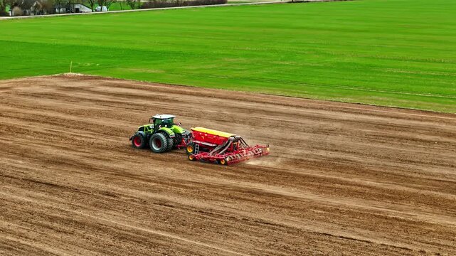Aerial view of a green tractor pulling a red seeder across a large brown ploughed field with vibrant green grass in the background in Riga, Latvia.