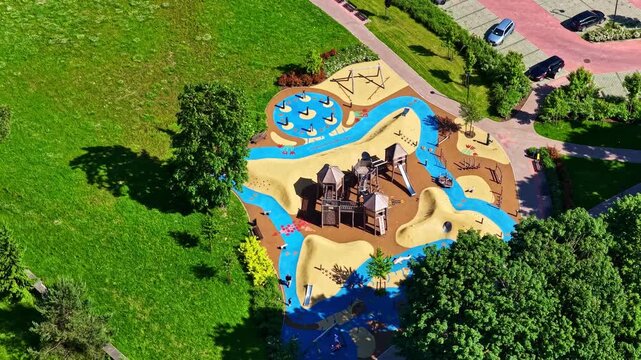 Aerial view of a colorful playground with wooden structures and blue rubber flooring surrounded by green grass and trees in Riga, Latvia.