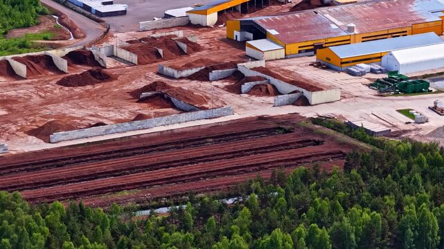 Aerial view of an industrial peat processing facility with large piles of dark brown material and storage buildings near a forest in Riga, Latvia.
