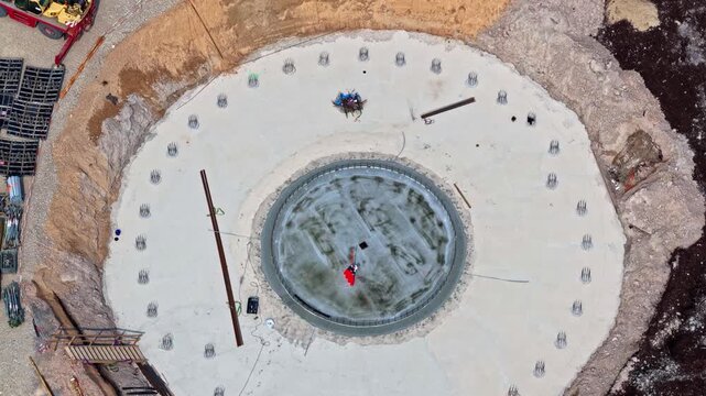 Aerial view of a circular concrete wind turbine foundation under construction with exposed rebar and surrounding earthworks in Riga, Latvia.