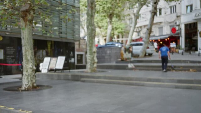 Blurred urban plaza scene with shallow bokeh focus on pavement, trees and out of focus storefronts and steps; backplate template overlay calm.