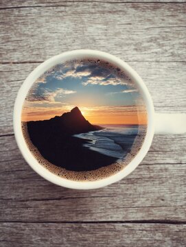 Coffee cup displaying mountain beach sunset landscape on wooden table top view creative concept