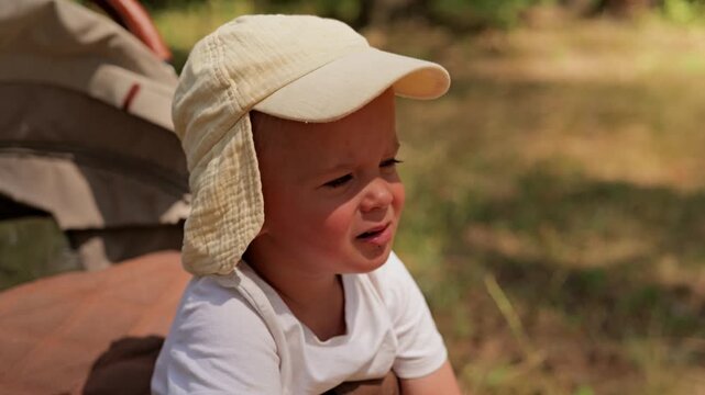 A young child sits on a blanket in a forest. The child wears a hat and has a serious expression. Sunlight shines through the trees around them