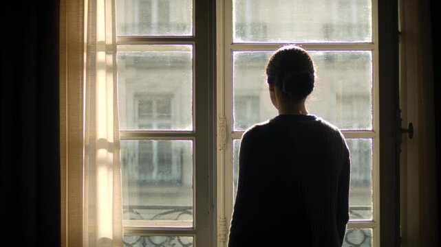 Sunlight drawing woman shifting toward window in flat, in dark sweater, sheer curtain, iron railing