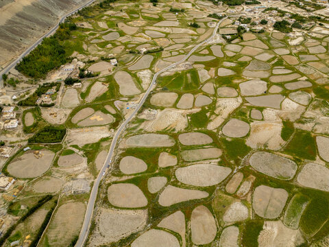 Aerial view of the agricultural landscape featuring irregular patch-like fields with green and brown textures, winding roads, and small village settlements in Zanskar, Ladakh, India.