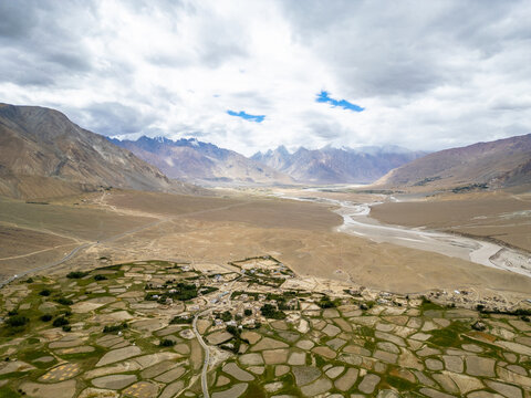 Aerial view of the Zanskar valley featuring patchwork agricultural fields, a winding river, and dramatic mountain ranges under a cloudy sky in Zanskar, Ladakh, India.