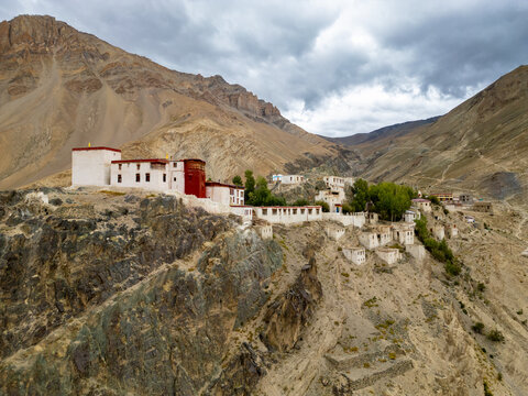Aerial view of the Zanskar monastery perched on a rugged cliffside with white buildings and red accents, surrounded by dramatic mountains under a cloudy sky in Zanskar, Ladakh, India.