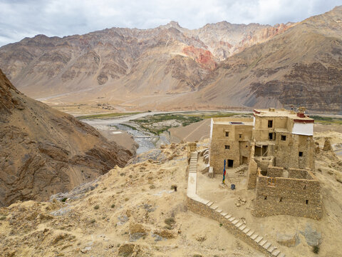 Aerial view of an ancient stone monastery perched on a rocky cliff overlooking the Zanskar valley with rugged mountains and a winding river in Zanskar, Ladakh, India.