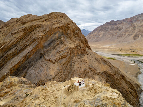 Aerial view of a remote Buddhist monastery perched on a rocky cliffside overlooking a winding river valley in the rugged Himalayan landscape of Zanskar, Ladakh, India.