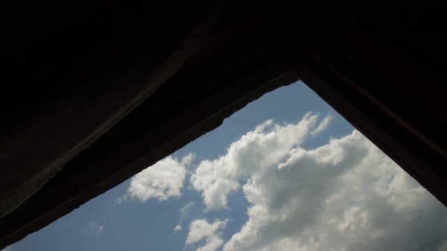 Time-lapse of white clouds moving across blue sky viewed through an open window with dark curtains