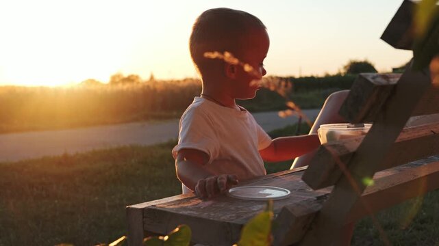 A child is at a wooden bench in an open field during sunset. The child focuses on a container, enjoying the moment as the sun sets in the background