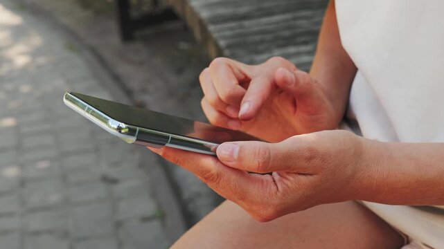 A person is sitting on a wooden bench in a park, looking at a smartphone. The sun is shining, and it is a warm afternoon