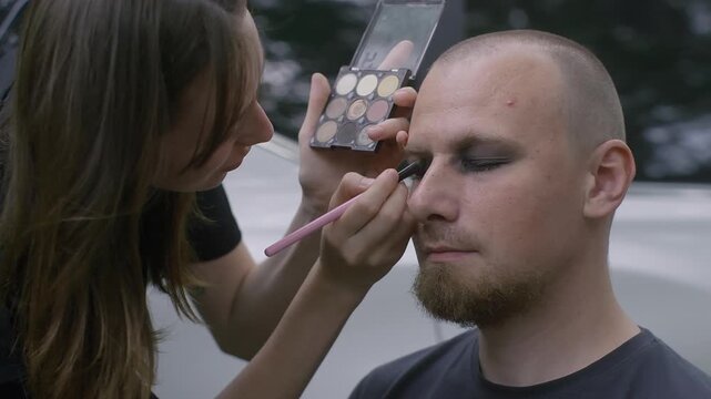 Professional makeup artist applies goth style cosmetics to male rock band member before concert performance, using eyeshadow palette, close up of face.