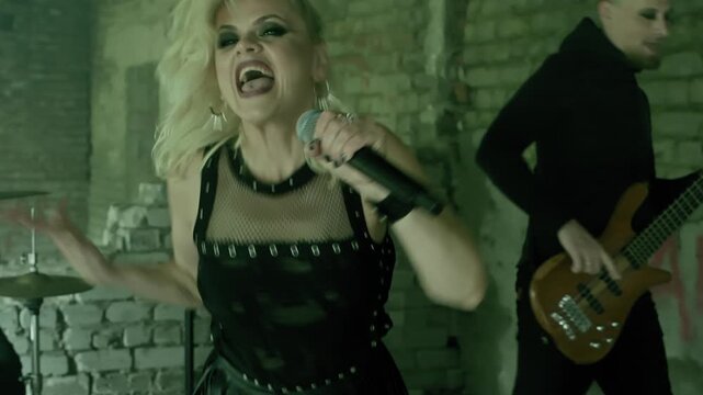 Female singer with blonde hair passionately performs with microphone while male guitarist plays bass in grungy, deteriorated brick interior space.