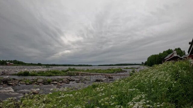 Wide landscape of fast rushing river rapids with rustic wooden building and summer flowers