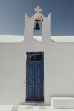 A small whitewashed chapel in Santorini, Greece, with a vivid blue door, bell, and cross &mdash; a serene example of Cycladic architecture and Mediterranean heritage.