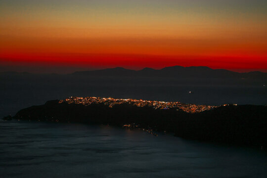 Twilight falls over Santorini, Greece, as Oia city lights shimmer along the coast beneath a fiery red and orange sky.