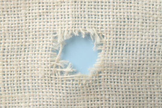 Burlap fabric with torn hole on light blue background, top view