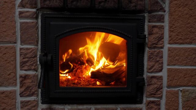 Burning firewood in a home fireplace with a closed glass door and brick frame. Cozy atmosphere with bright orange flames and glowing embers inside a wood stove, winter heating concept