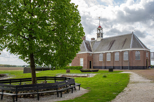 Schokland, Netherlands - 17 May 2015: View of the historic Enkhuizerkerk church on the former island of Schokland with a large green tree and circular bench under a cloudy sky.