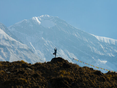 Aerial view of a person performing a yoga tree pose on a grassy hill with prayer flags against a backdrop of snow-capped mountains in Nepal.