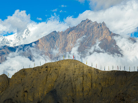 Aerial view of a rocky mountain ridge adorned with colorful prayer flags, set against a backdrop of towering snow-capped Himalayan peaks and drifting clouds in Nepal.