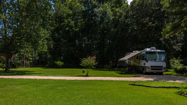 A large RV is parked on a grassy area surrounded by lush green trees. The scene is peaceful with ample sunshine and clear skies.