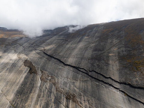Aerial view of a steep, dark grey mountain rock face with prominent geological layers and deep fissures under a cloudy sky in Nepal.