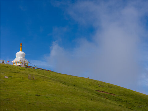 Aerial view of a white Buddhist stupa perched on a lush green hillside adorned with colorful prayer flags under a bright blue sky in Nepal.