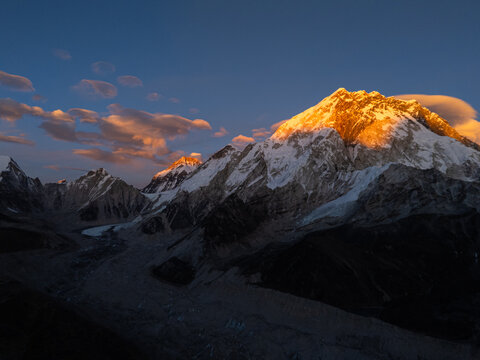 Aerial view of the Himalayan mountain range at golden hour with snow-capped peaks glowing in the sunset light, deep shadows in the valleys, and wispy clouds in the blue sky, Nepal.
