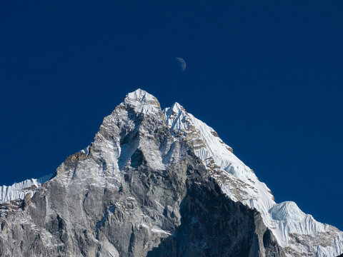 Aerial view of a snow-capped Himalayan mountain peak with jagged rock formations and a crescent moon visible in the clear deep blue sky in Nepal.