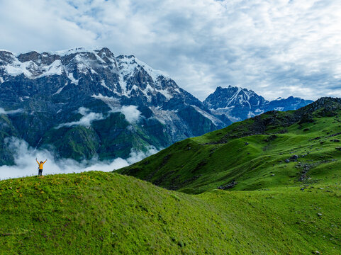 Aerial view of a hiker in a yellow jacket standing with arms raised on a lush green mountain ridge overlooking the snow-capped Himalayan peaks and clouds in Nepal.