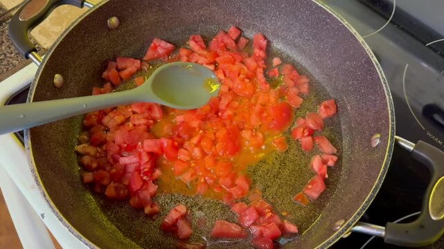 Chopped tomatoes are sauteing in a frying pan with a gray spoon, showcasing the cooking process in a kitchen setting, with visible stove and countertop elements enhancing the scene