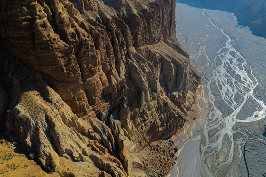 Aerial view of a massive, rugged mountain cliff face towering over a wide, braided riverbed in the arid landscape of Nepal.