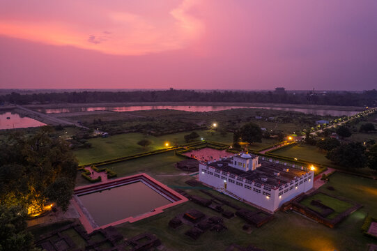 Aerial view of the Maya Devi Temple in Lumbini, featuring the sacred pond, ancient ruins, and illuminated temple structure under a vibrant sunset sky, Nepal.