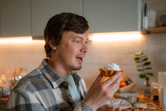 Man with a strong sweet tooth holding a small cupcake topped with whipped cream, expressing a craving for sugar and anticipating the satisfying taste of the dessert within a home kitchen