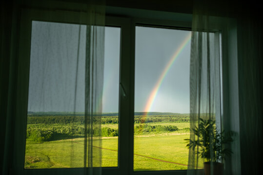 Window framing a vibrant colorful rainbow arching across a green summer field and distant forest, offering hope and a fresh perspective on nature's beauty from inside a home